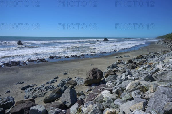 Rocks spread along a sunny sandy beach with waves in the background, West Coast, South Island, New Zealand