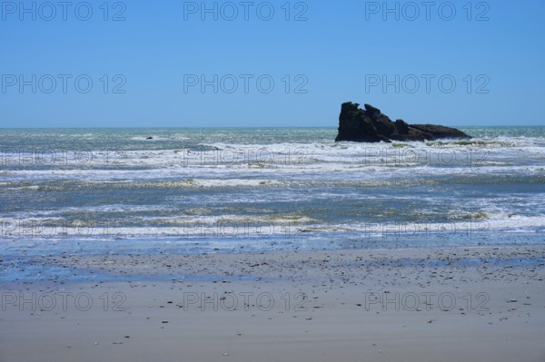 The picture shows a beach with rocks and gentle waves under a clear blue sky, West Coast, South Island, New Zealand