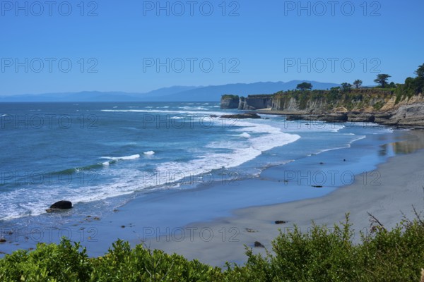 A peaceful coastal landscape with blue waves crashing against sand and cliffs under a clear blue sky, Cape Foulwind, West Coast, South Island, New Zealand