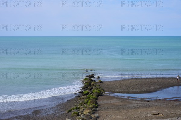 View of the calm sea with rocks leading into the water and a lone walker on the beach, Mana Bay, Riverview Lookout, Patea, Taranaki, North Island, New Zealand