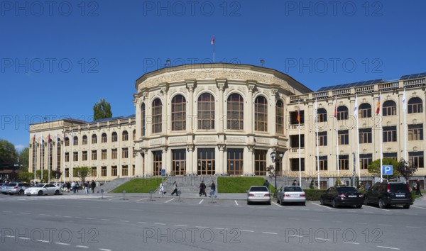 A large classical building with flags, cars and people under a clear sky, Gyumri, Gyumri, Gyumri, Shirak province, Armenia