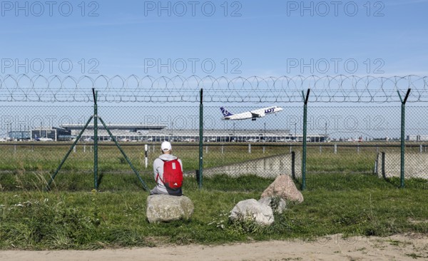 A man sits on a stone and watches an aircraft of the Polish airline LOT take off through a barbed wire fence at BER Airport, Berlin Schönefeld, 28/09/2025, Schönefeld, Brandenburg, Germany