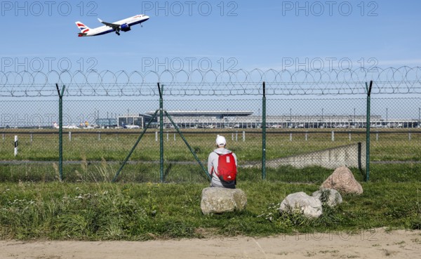 A man sits on a stone and watches the take-off of a British Airways plane through a barbed wire fence at BER Airport, Berlin Schönefeld, 28/09/2025, Schönefeld, Brandenburg, Germany