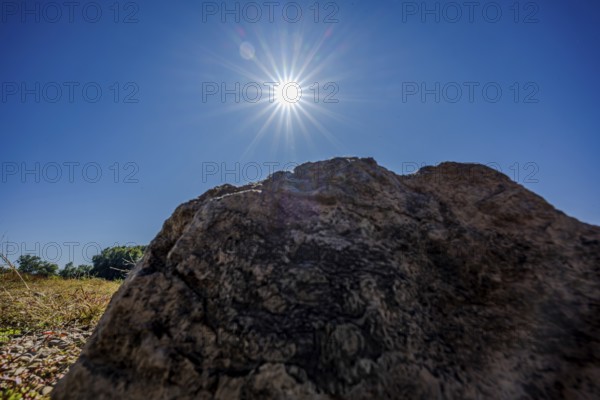 A large stone stands in the foreground under a bright blue sky. The sun shines brightly and creates a warm atmosphere in the surroundings. Bogen, Lower Bavaria