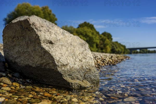 A large stone lies on the bank of a calm river. The water is clear and reflects the blue sky. Trees line the bank and it is a beautiful, sunny day. Bogen, Lower Bavaria