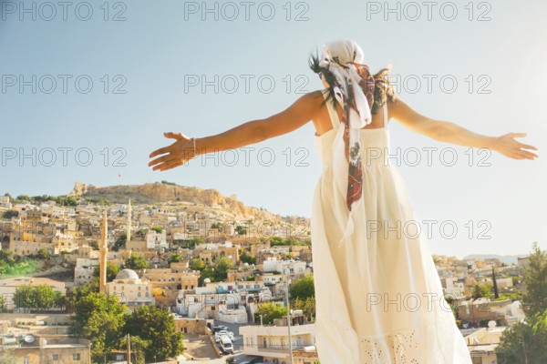 Traveler woman in white dress arms outstretched looking over panoramic view of historic city of Mardin Turkey with mosque minarets and traditional houses and Mardin castle on hilltop