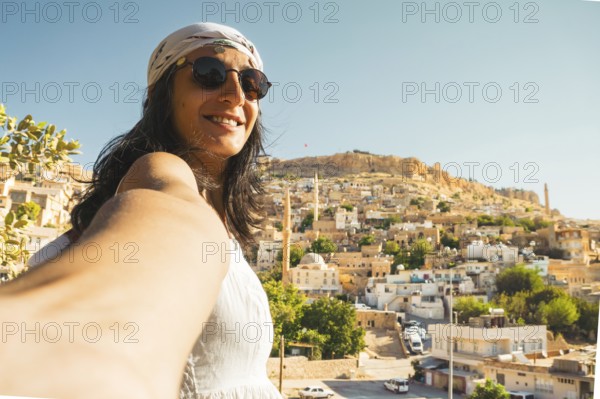 Smiling woman in sunglasses taking selfie with panoramic skyline of historic Mardin Turkey and mosque minarets on Mesopotamian hillside