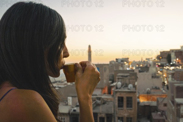 Tourist woman enjoying traditional Mirra coffee on rooftop with Mardin skyline and Mesopotamian plains at sunset, traditional Turkish Arab culture