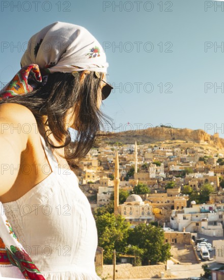 Traveler woman in white dress arms taking selfie self-portrait ooking over panoramic view of historic city of Mardin Turkey with mosque minarets and traditional houses and Mardin castle on hilltop