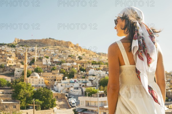 Traveler woman in white dress looking over panoramic view of historic city of Mardin Turkey with mosque minarets and traditional houses and Mardin castle on hilltop