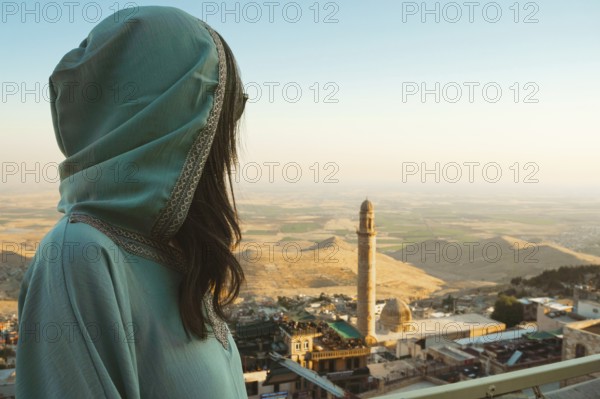Smiling woman traveler in abaya standing above Mardin Turkey with panoramic view of Mesopotamian plains and historic mosque minaret at sunset
