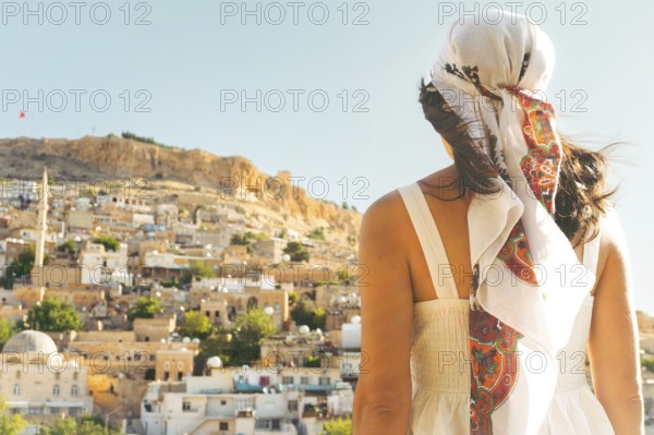Traveler woman in white dress looking over panoramic view of historic city of Mardin Turkey with mosque minarets and traditional houses and Mardin castle on hilltop
