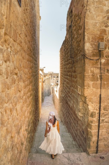 Traveler woman in white dress walking through narrow stone alley in historic city of Mardin Turkey with traditional architecture