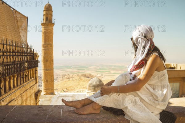 Traveler woman in white dress sitting near Great Mosque of Mardin Ulu Camii with panoramic view of Mesopotamian plains and historic minaret