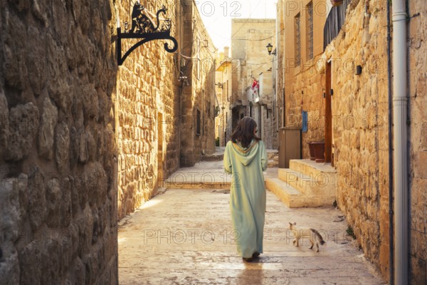 Woman in traditional abaya walking with cat through narrow stone alley in historic city of Mardin Turkey with ancient architecture