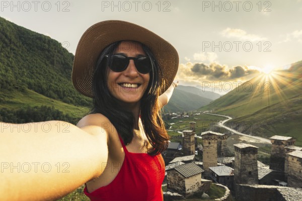 Smiling woman in hat and red dress taking selfie at sunset with medieval towers of Ushguli Georgia in Caucasus mountains