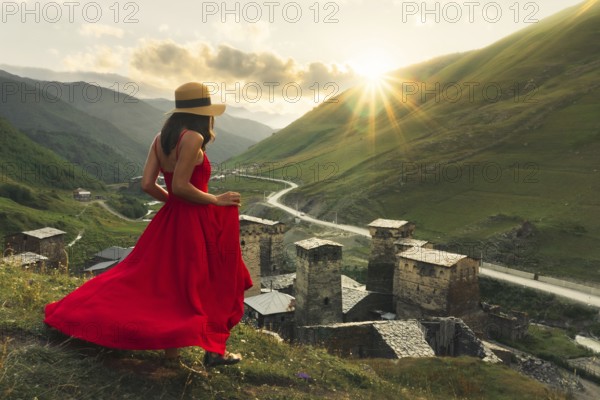 Woman in red dress looking at medieval towers of Ushguli Georgia during sunset with mountains and Enguri valley landscape