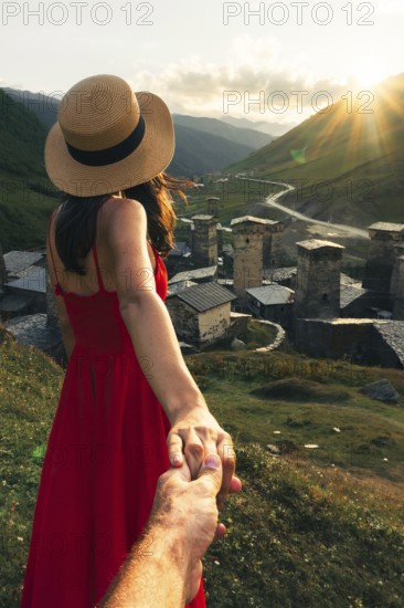Young Couple holding hands leading way in Ushguli Georgia woman in red dress leading towards medieval towers at sunset in Caucasus mountains