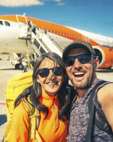 Cheerful caucasian couple taking selfie in front of passenger airplane before boarding flight symbolizing travel, tourism, adventure, and vacation lifestyle