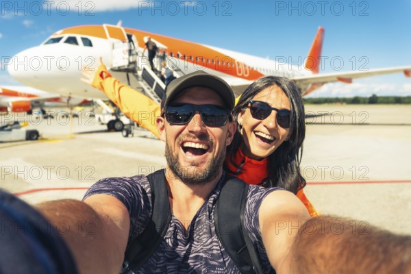 Cheerful caucasian couple taking selfie in front of passenger airplane before boarding flight symbolizing travel, tourism, adventure, and vacation lifestyle
