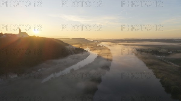 In the early morning, thick fog lies over the river. The sun slowly rises and illuminates the landscape. A quiet, picturesque moment in nature. Bogen, Lower Bavaria