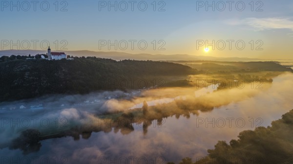 The sunrise illuminates the landscape over the calm Danube. A light mist floats gently over the water, while the mountains are clearly visible in the background. Bogen, Lower Bavaria