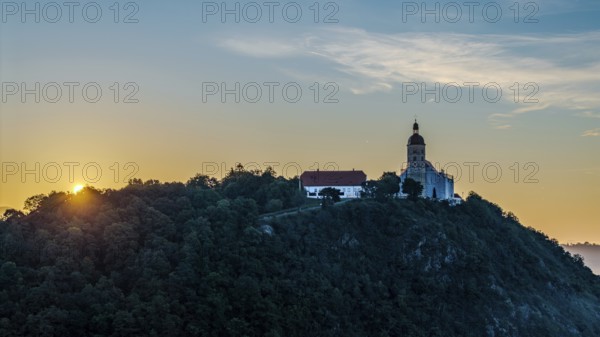 The sunrise illuminates the pilgrimage church of the Assumption of the Virgin Mary on the Bogenberg. The sky is tinged with soft colours. Trees surround the site and create a peaceful atmosphere. Bogen, Lower Bavaria