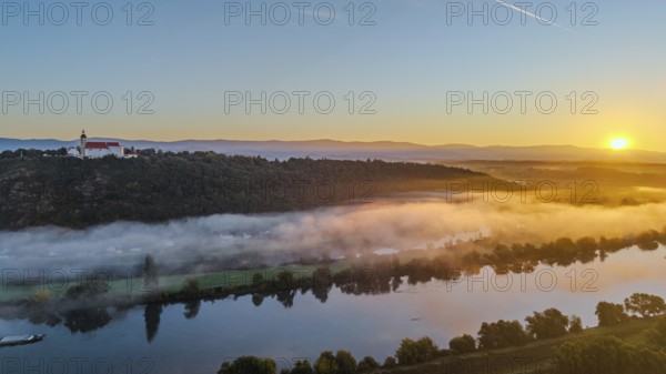 The sunrise bathes the Danube in warm light. A light mist hovers over the water, while the pilgrimage church of the Assumption of the Virgin Mary sits majestically on the Bogenberg. A peaceful morning atmosphere. Bogen, Lower Bavaria