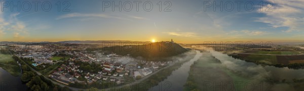 The sun rises over a city surrounded by fog. The picture shows the river landscape of the Danube and the Bogenberg with the historic pilgrimage church of the Assumption of the Virgin Mary in the background. Bogen, Lower Bavaria