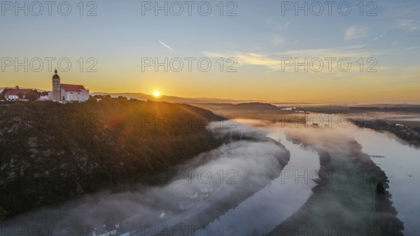 The sun rises over the Danube while mist drifts over the water. A tranquil landscape with trees and the historic pilgrimage church of the Assumption of the Virgin Mary on Bogenberg hill stretches out in the foreground. Bogen, Lower Bavaria