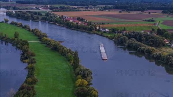 A cargo ship sails quietly on the Danube. The surroundings are green with fields and trees. The day ends and the sky shows soft colours. Bogen, Lower Bavaria