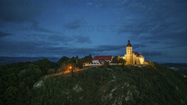 The pilgrimage church of the Assumption of the Virgin Mary stands majestically on the Bogenberg, while the evening sky glows in soft colours. The orange-coloured lighting lends the scene a tranquil atmosphere. Bogen, Lower Bavaria