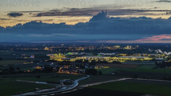 The scene shows an expansive landscape at dusk. Illuminated buildings and streets are visible in the distance, surrounded by clouds and rolling hills. Straubing, Lower Bavaria