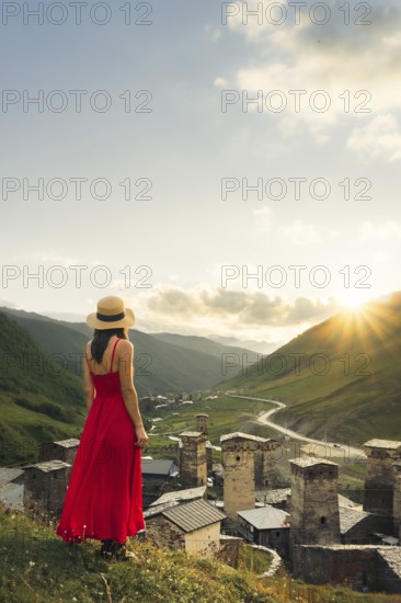 Young pretty Woman traveler in red dress looking at medieval towers of Ushguli Georgia during sunset with mountains and Enguri valley landscape
