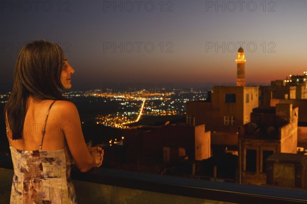 Tourist woman on terrace restaurant balcony watching illuminated skyline of Mardin with glowing minaret and Mesopotamian plains at dusk, cultural travel scene