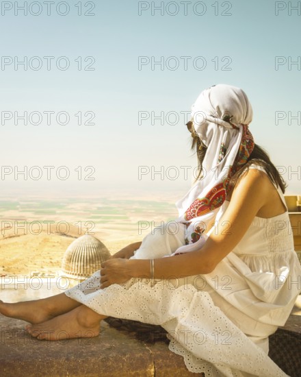 Traveler woman or tourist wear white dress sitting near Great Mosque of Mardin overlooking vast Mesopotamian plains with historic architecture in foreground