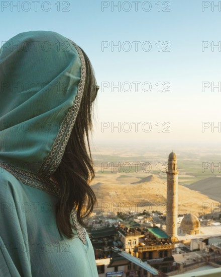 Smiling woman traveler in abaya standing above Mardin Turkey with panoramic view of Mesopotamian plains and historic mosque minaret at sunset