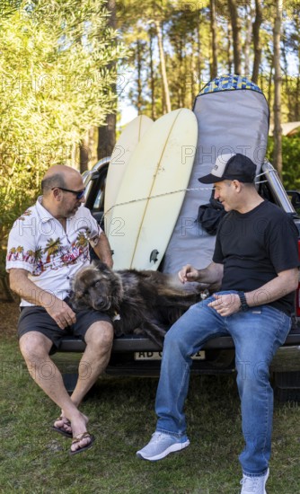 Two male friends talking sitting on a truck with their dog and surfboards