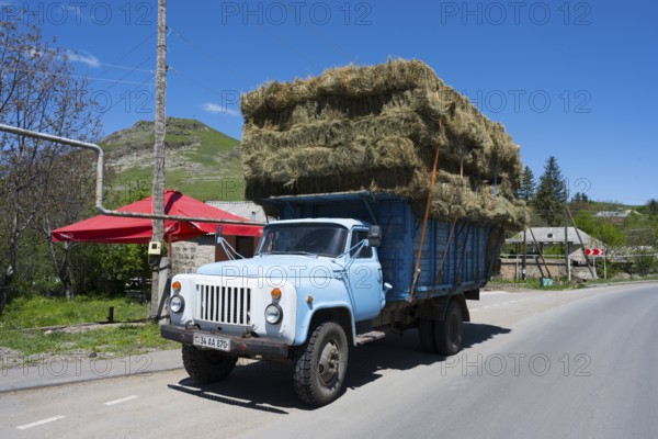 A lorry drives along a village road, loaded with many layers of hay bales, surrounded by green hills, Armenia