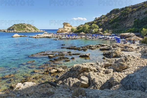 Grande Blue rocky beach with blue sea, umbrellas and surrounding cliffs under clear skies, Grande Blue Beach, Stegna, Archangelos, Rhodes Island, Greece