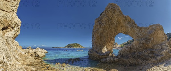 Stone arch on Grande Blue Beach in front of a small island and a rocky coastline under a clear blue sky, Grande Blue Beach, Stegna, Archangelos, Rhodes Island, Greece