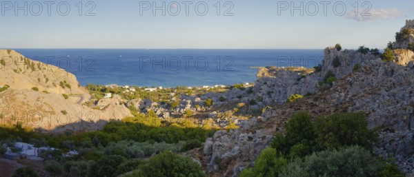 Coastal landscape of Stgna overlooking the sea, framed by rocky hills and lush vegetation, under a blue sky, Grande Blue Beach, Stegna, Archangelos, Rhodes Island, Greece