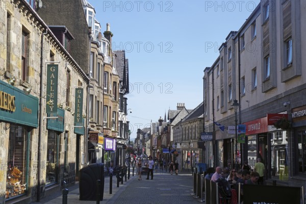 Busy shopping street with people and traditional buildings under a blue sky, Fort William, Scotland, United Kingdom