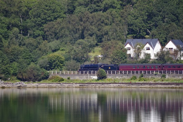 A train travelling along a loch, past houses and a dense green forest, Hogwarts Express, Jacobite Steam Train, Loch Eil, Scotland, Great Britain