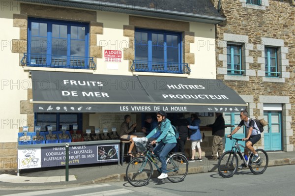 Cyclists on the Quai John Kennedy pass a shop selling seafood such as oysters and mussels, Cancale, Brittany, France