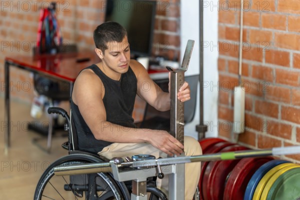 Young man living with a disability sitting in a wheelchair while adjusting a barbell rack during a fitness workout session at an accessible gym, showing dedication