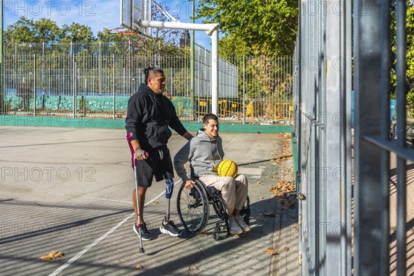 Friends celebrating sports inclusion on a sunny outdoor basketball court, one man with crutches and a prosthetic leg stands while pushing his smiling friend in a wheelchair holding a yellow ball