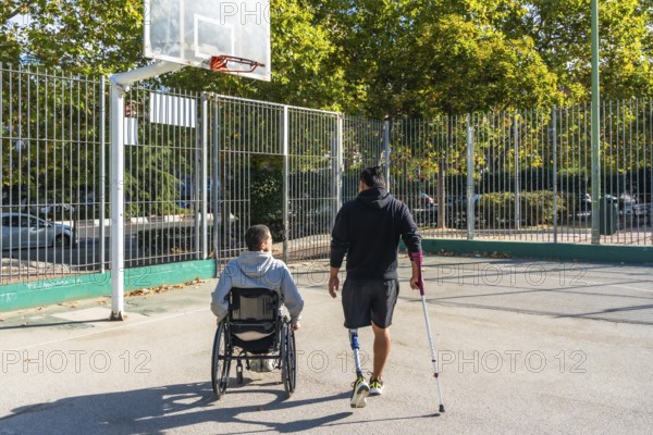Two young men, one in a wheelchair and the other with a prosthetic leg, are walking together towards a basketball hoop on an outdoor court, symbolizing inclusion and adaptive sports