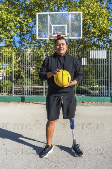 Man with prosthetic leg stands on outdoor basketball court holding a yellow ball, exuding determination, fitness and inclusive athleticism in an urban park setting