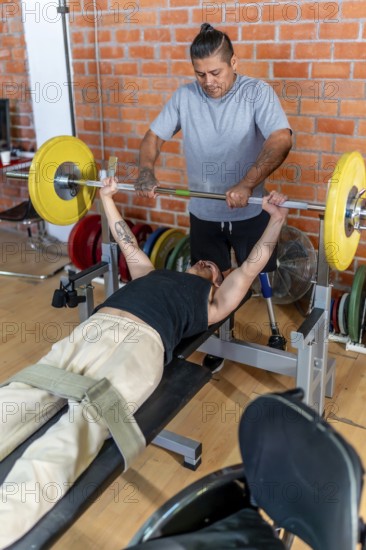Young man with a prosthetic leg performing bench press exercises at the gym, receiving support and spotting from his personal trainer, focusing on strength and rehabilitation